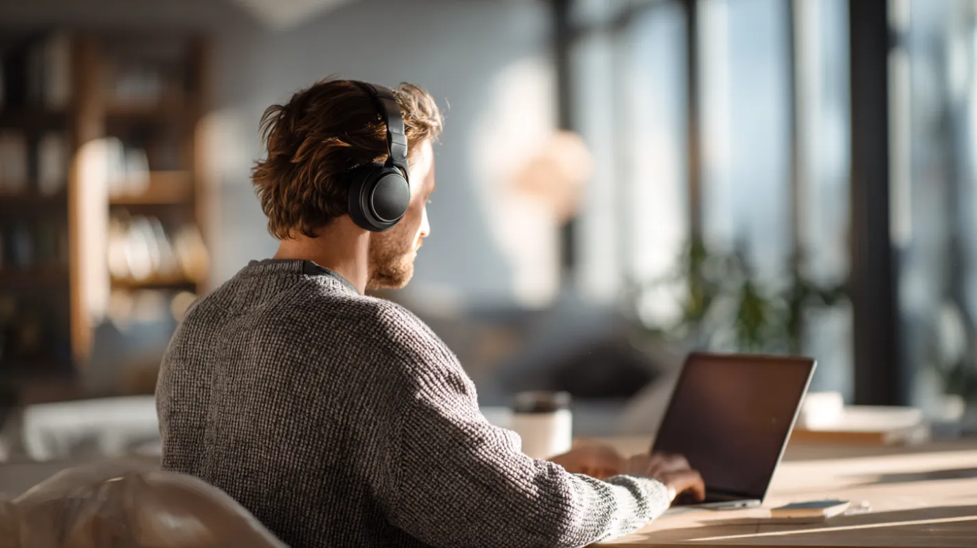Man wearing headphones working on laptop at desk in bright modern office with city view.