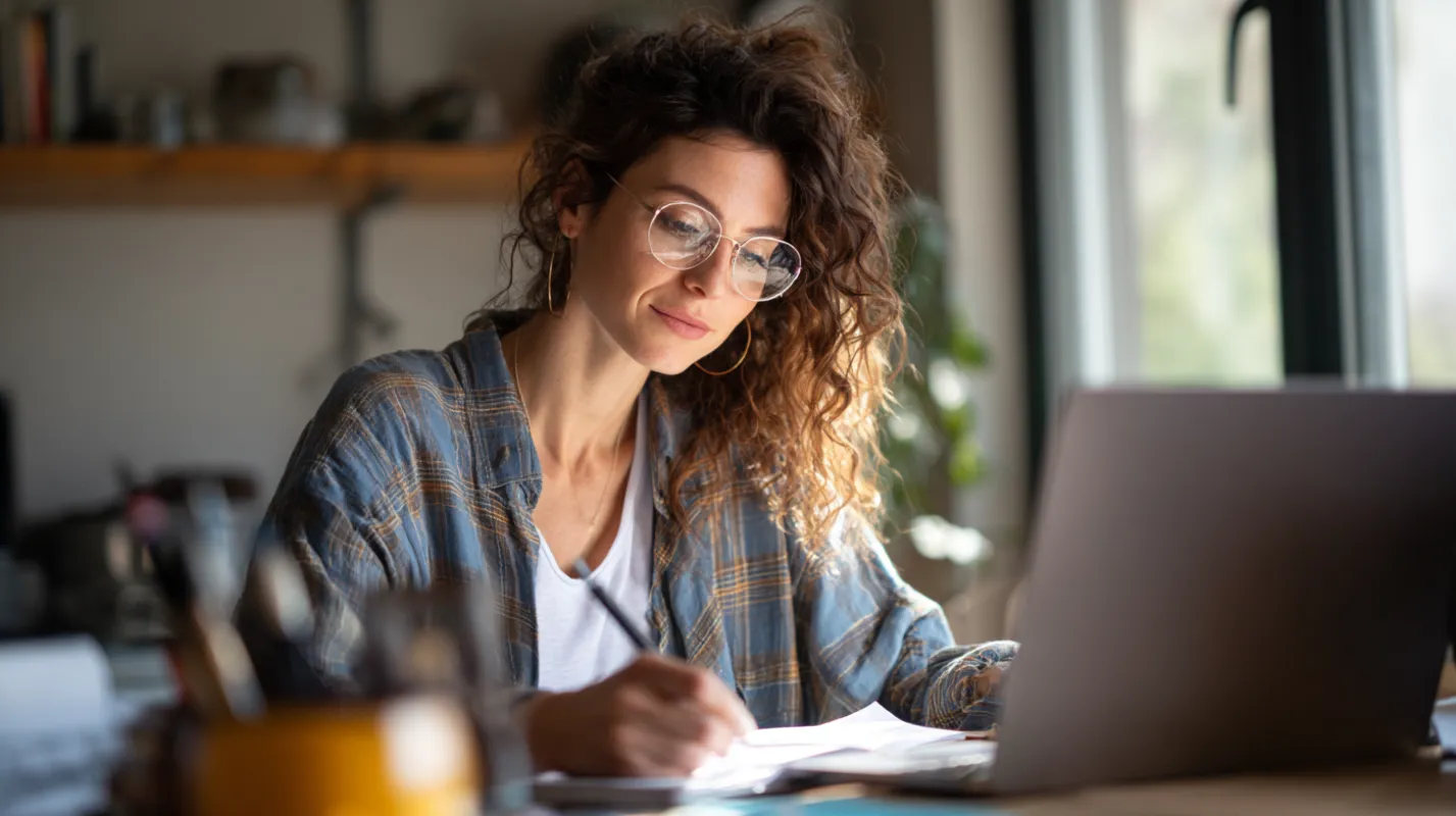 Woman with curly hair and glasses working at laptop while writing notes at wooden desk in bright office space.
