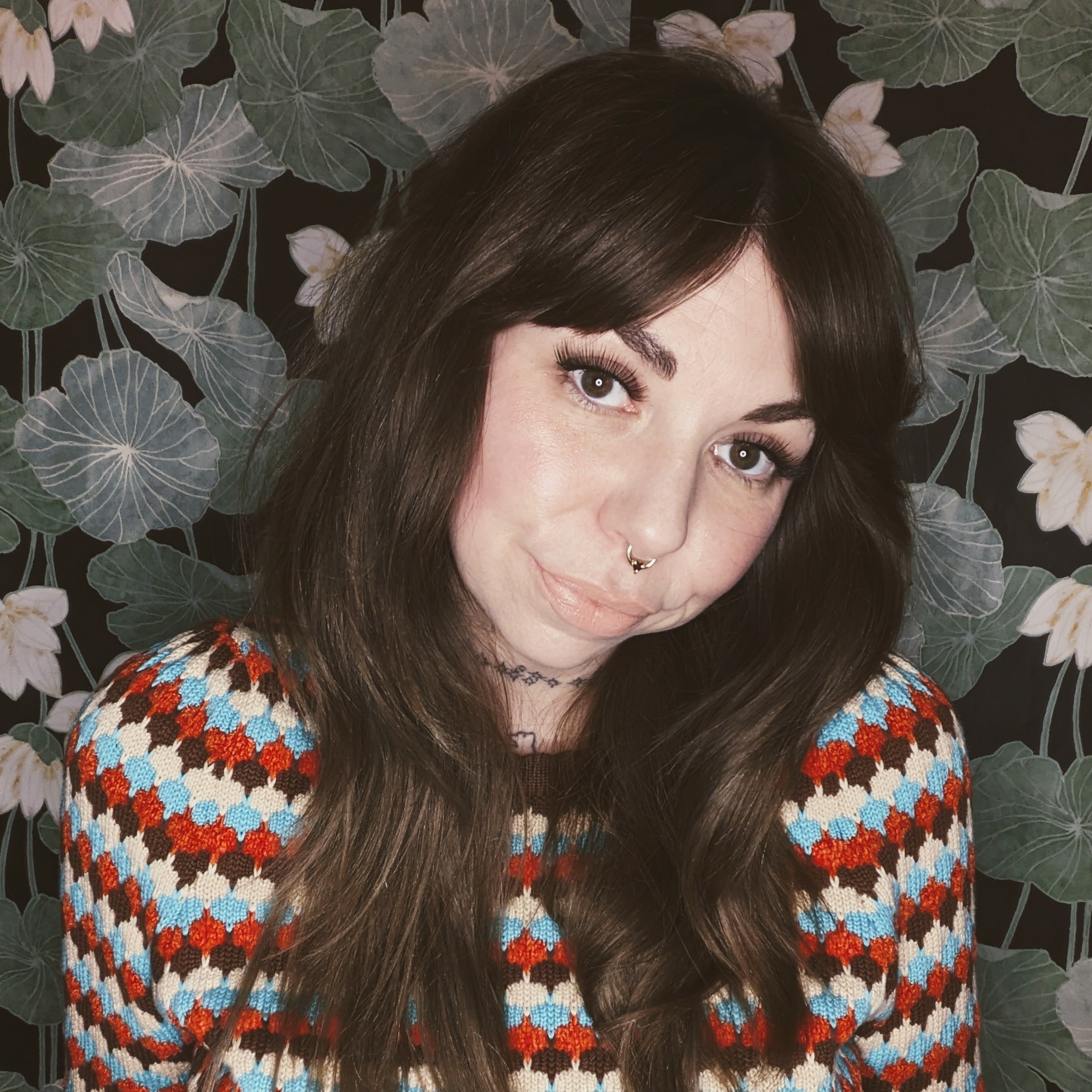 Picture of Annie with dark hair and nose piercing wearing a red, blue, and cream patterned sweater against a floral wallpaper background.