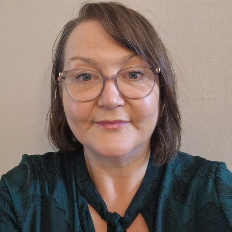 Woman with shoulder-length brown hair wearing clear-framed glasses and a dark teal patterned shirt against a neutral beige background.