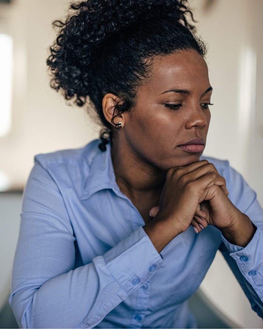 Woman in light blue shirt sitting thoughtfully with hand to chin, looking downward in contemplative pose.